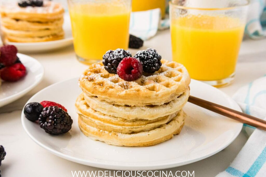 Four vegan waffles topped with berries on a white plate, with two cups of orange juice in the background