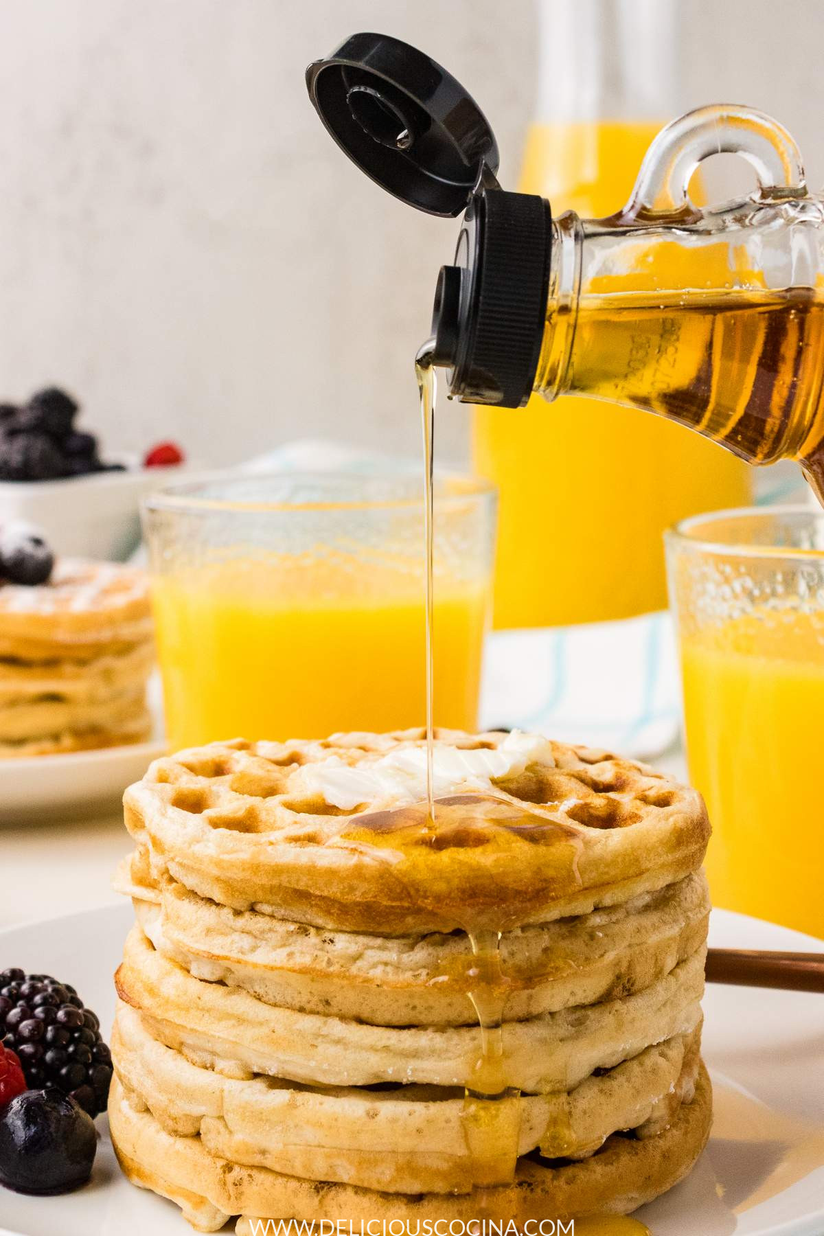 Four waffles with syrup being pour on top, with two cups and a pitcher of orange juice in the background